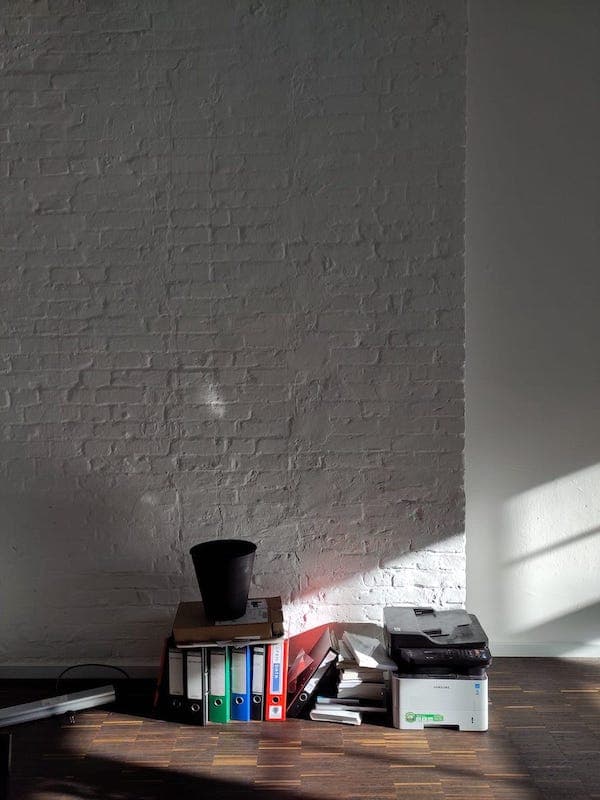 a white brick wall with binders and a printer on the wooden floor in front of it.